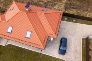 Aerial top view of house metal shingle roof with attic windows and black car on paved yard.